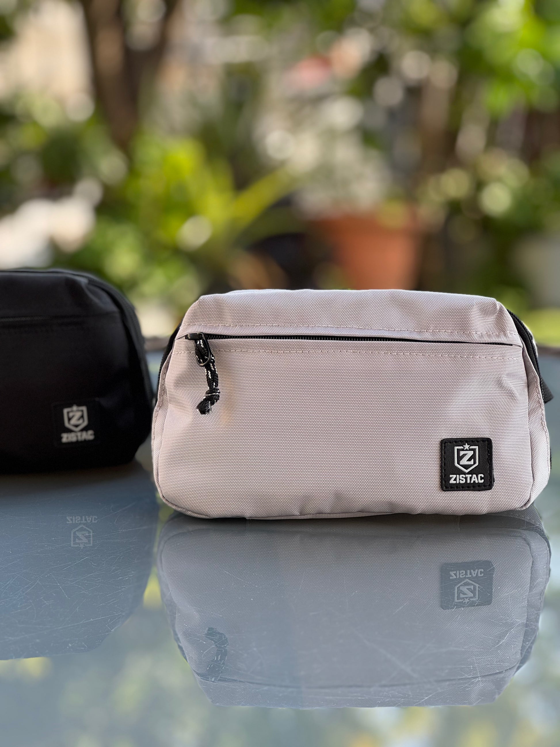 Two bags, one black and one beige, on a reflective surface with a blurred green outdoor background.