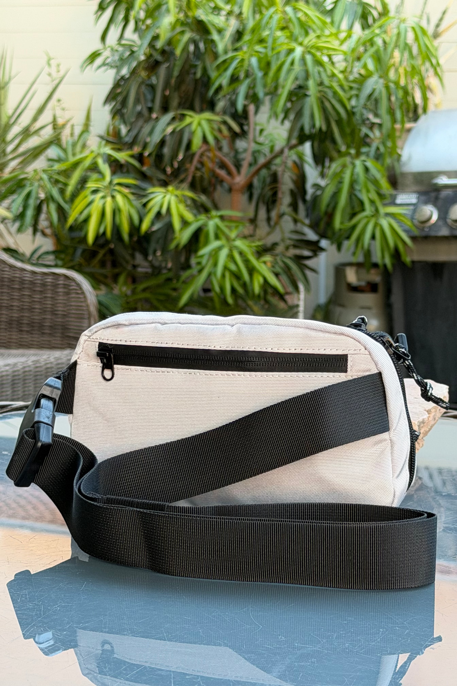 A beige canvas crossbody bag with a black webbing strap, resting on a glass table in an outdoor patio setting with green plants in the background.